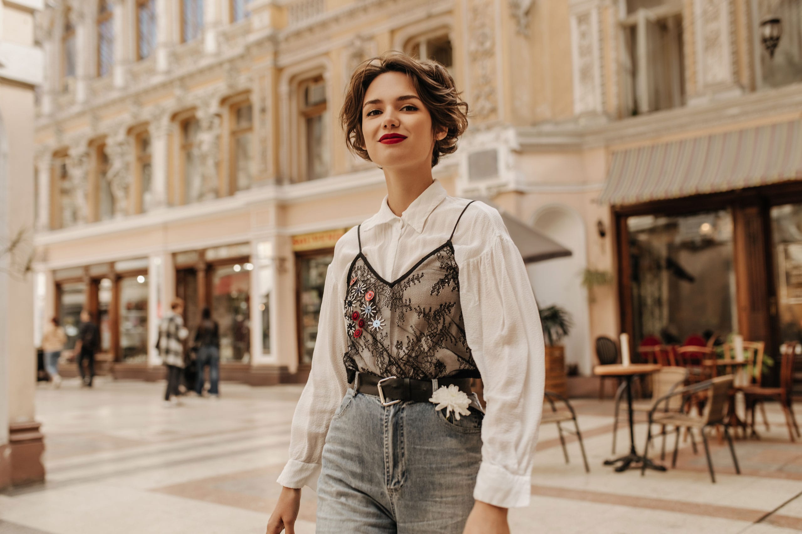 Junge Frau mit kurzen Haaren und rotem Lippenstift in einer historischen Einkaufspassage mit verzierten Fassaden. Sie trägt eine weiße Bluse unter einem schwarzen Spitzen-Top und eine Jeans mit Gürtel.